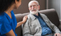 Young friendly african female caregiver talking chatting to happy senior man in hallway of nursing home. Picture of smiling nurse assisting senior elderly man