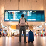 Asian male traveler with wheeled luggage checking for flight schedule on arrival and departure board at Kuala Lumpur International Airport